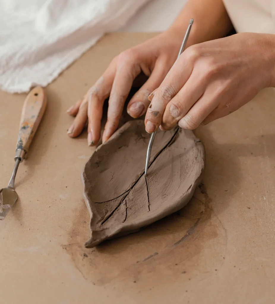 Gros plan sur des mains gravant les nervures d'une coupelle en forme de feuille en argile crue, avec des outils de céramiste sur l'établi
