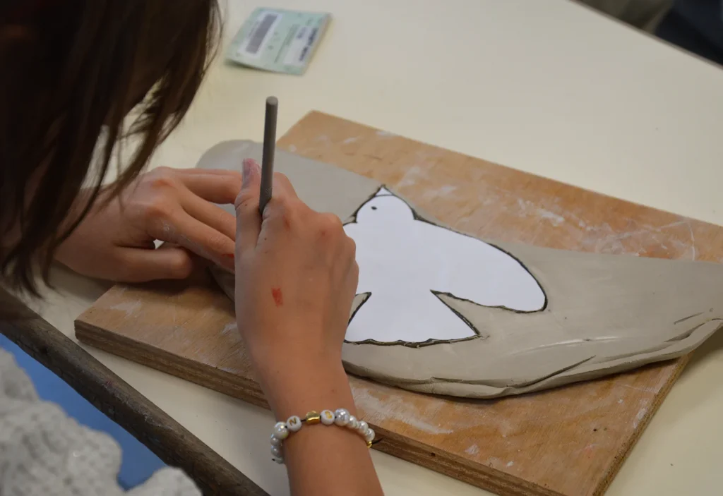 Enfant traçant le contour d'un gabarit oiseau en papier sur une plaque d'argile lors d'un atelier modelage à l'école d'Oms